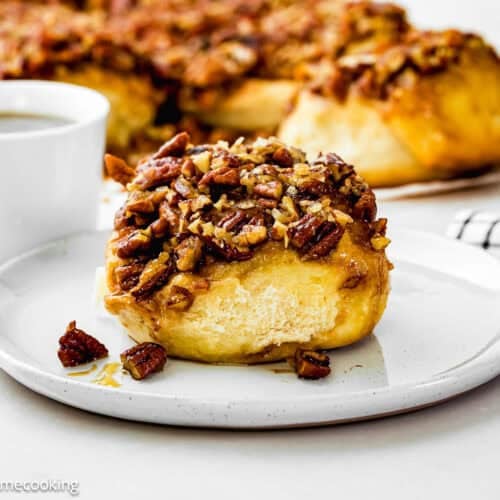 An Eggless Sticky Bun topped with chopped pecans sits on a white plate, accompanied by a cup of coffee and more buns in the background.
