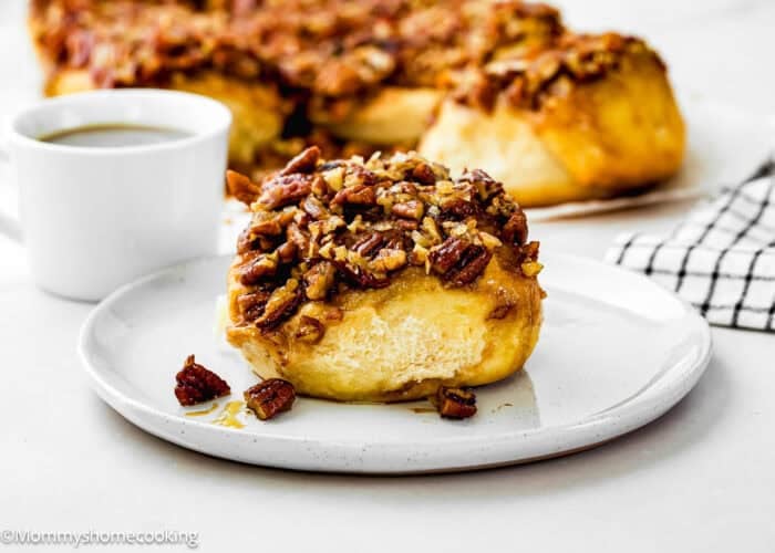 An Eggless Sticky Bun topped with chopped pecans sits on a white plate, accompanied by a cup of coffee and more buns in the background.