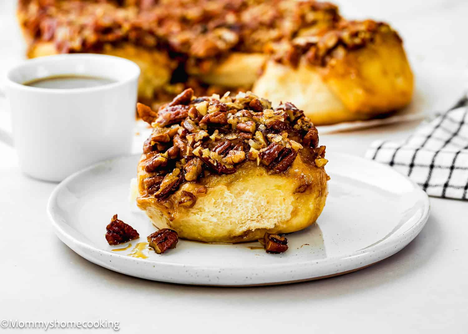 An Eggless Sticky Bun topped with chopped pecans sits on a white plate, accompanied by a cup of coffee and more buns in the background.