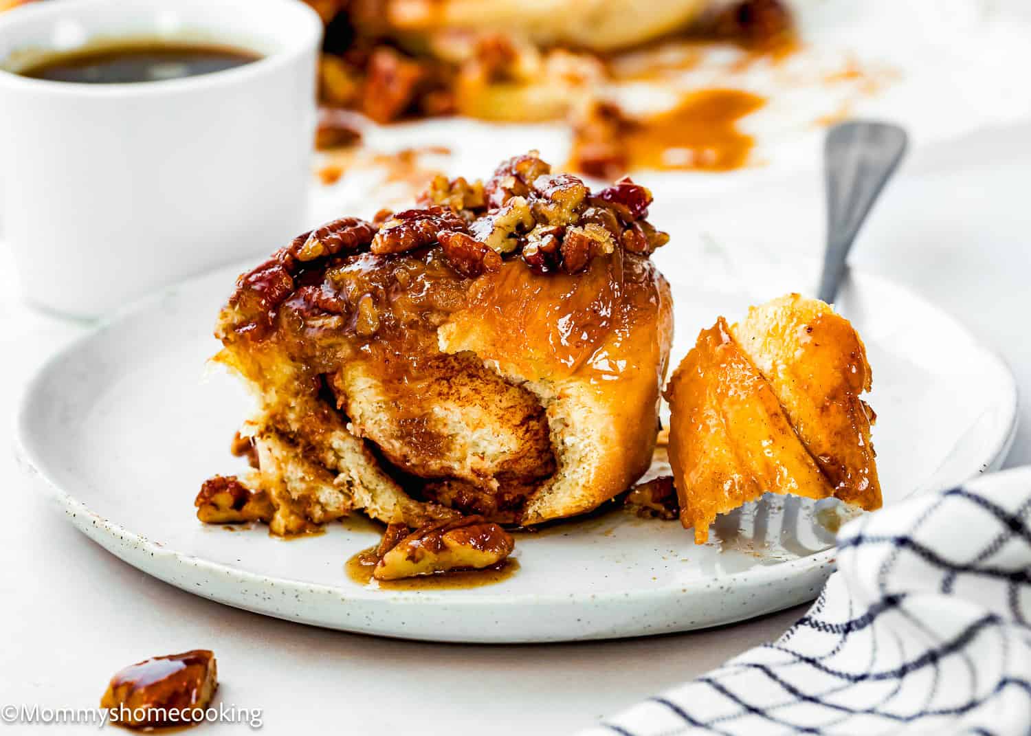 A partially eaten Eggless Sticky Bun topped with chopped pecans sits on a plate, with a fork holding a piece and a cup of coffee in the background.
