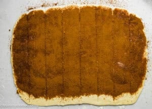 Rectangular sheet of dough topped with a layer of cinnamon sugar, cut into long strips on a floured surface—perfect for making Eggless Sticky Buns.