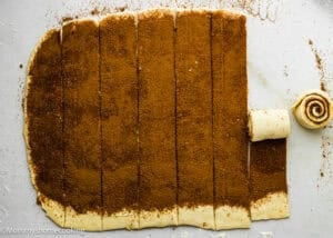 Rectangular sheet of dough covered with cinnamon sugar, sliced into strips; one strip is partially rolled into a spiral. Perfect for making Eggless Sticky Buns.