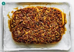 An eggless sticky bun, rectangular in shape and topped with a generous layer of chopped pecans, sits on parchment paper on a baking sheet.