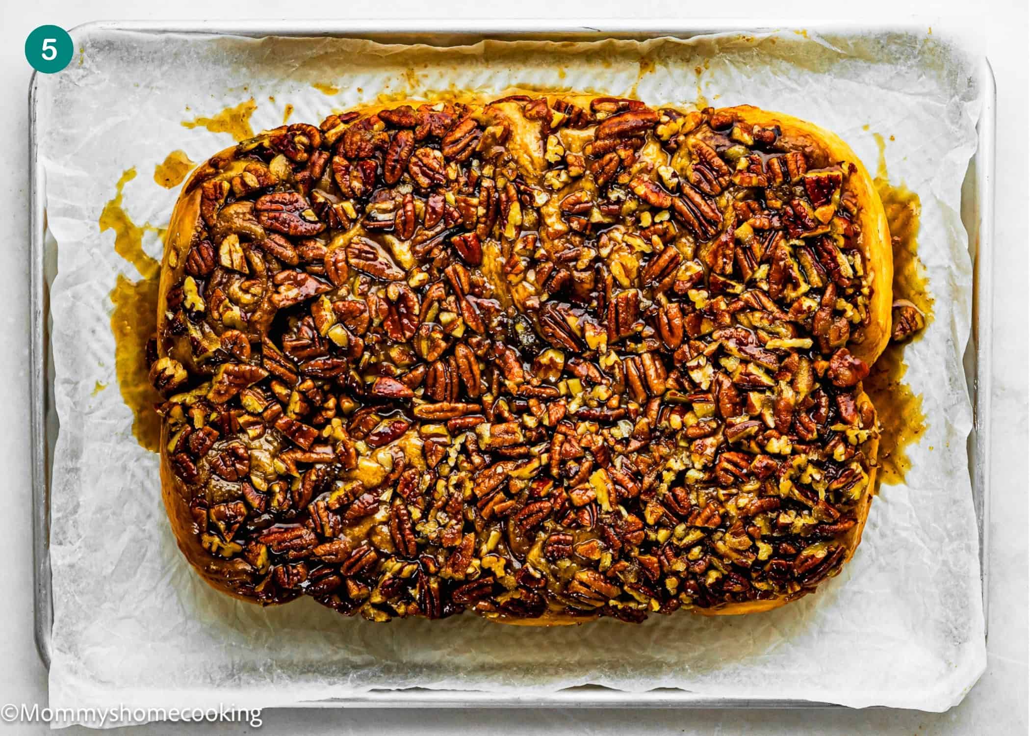 An eggless sticky bun, rectangular in shape and topped with a generous layer of chopped pecans, sits on parchment paper on a baking sheet.