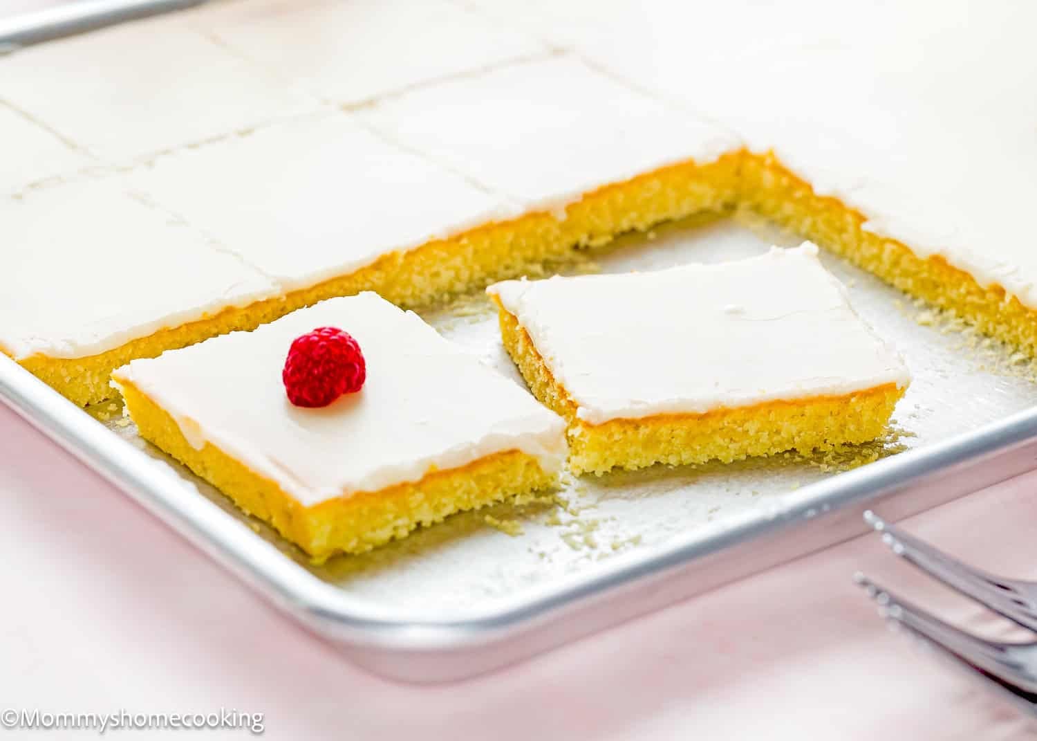 A tray of Eggless White Texas Sheet Cake bars with white frosting; one bar is topped with a raspberry and several pieces are cut and separated.