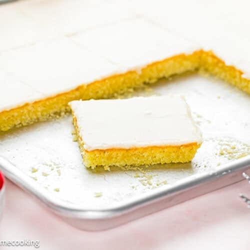 A rectangular Eggless White Texas Sheet Cake with one square piece removed rests on a metal baking pan. A small bowl of raspberries and two forks are visible in the foreground.