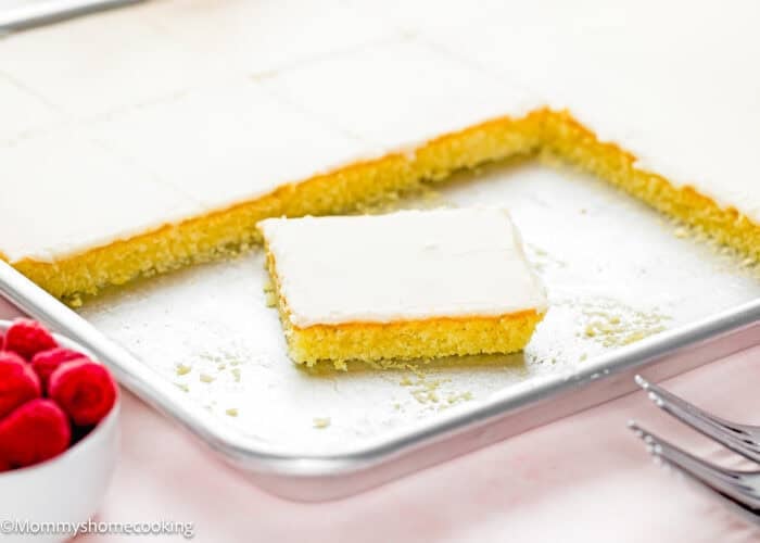 A rectangular Eggless White Texas Sheet Cake with one square piece removed rests on a metal baking pan. A small bowl of raspberries and two forks are visible in the foreground.