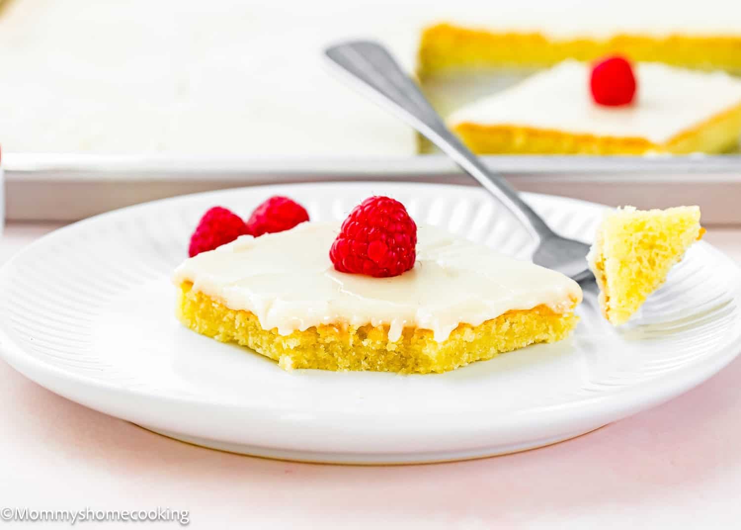 A square piece of frosted yellow cake, inspired by Eggless White Texas Sheet Cake, is topped with a raspberry and served on a white plate with a fork and two raspberries.