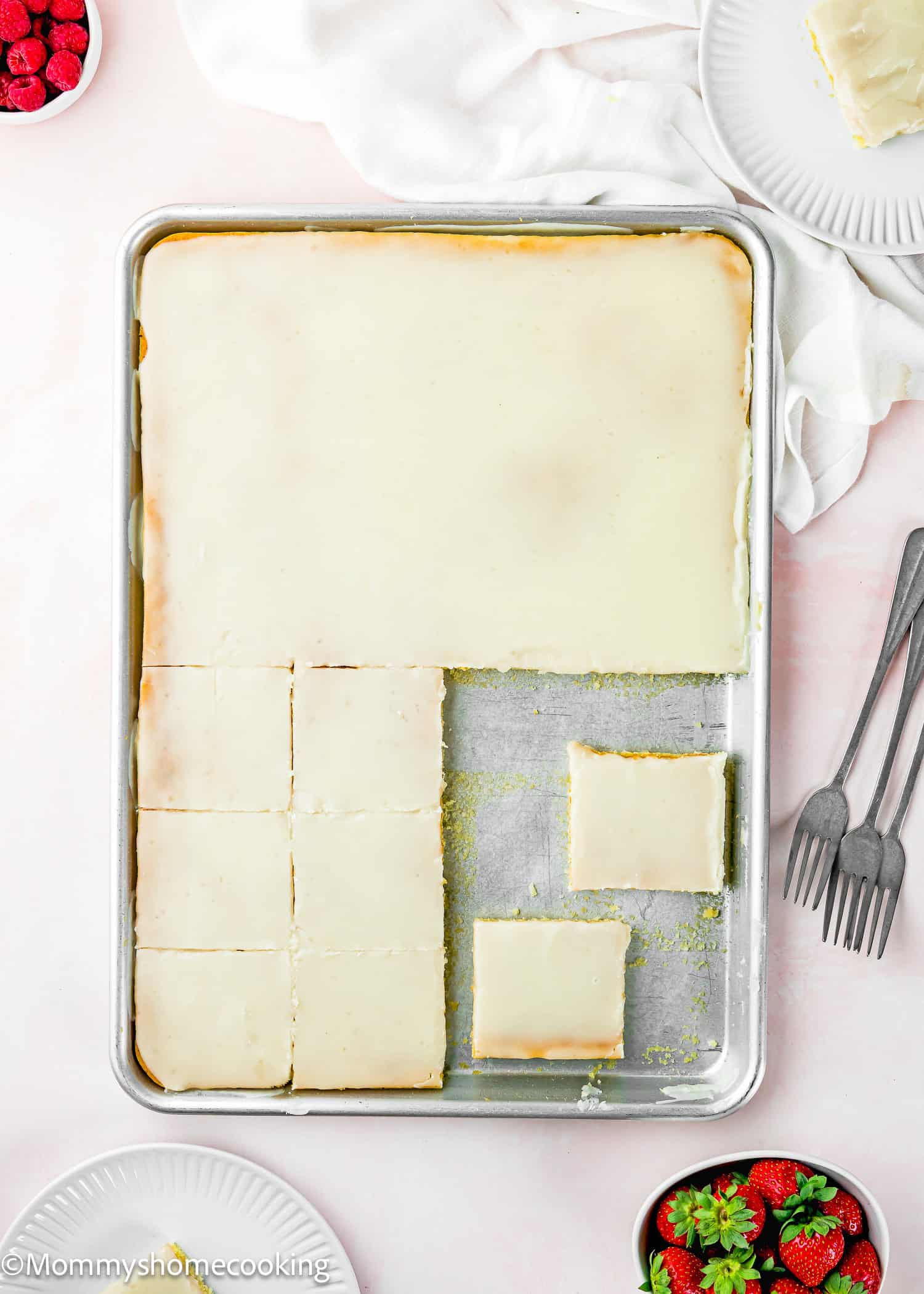 A sheet pan of frosted lemon bars with several bars cut and removed, surrounded by plates, forks, a bowl of strawberries, and an Eggless White Texas Sheet Cake on a light surface.