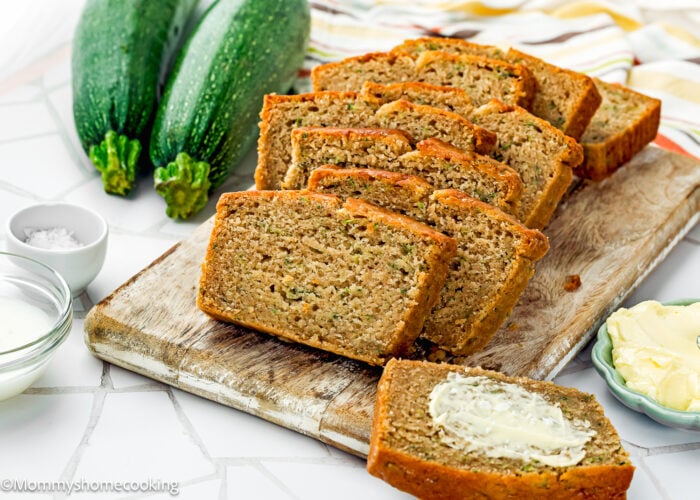Sliced zucchini bread is arranged on a wooden board, with butter on the side and fresh zucchinis in the background—perfect for showcasing this Eggless Zucchini Bread Recipe.