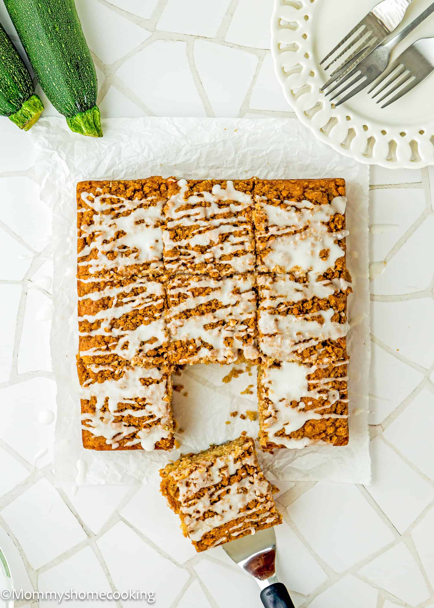 Iced oat cake with nine square pieces, one piece being served, on parchment paper beside a plate, forks, and fresh zucchini on a white tiled surface. This delicious Eggless Zucchini Coffee Cake is perfect for any occasion.