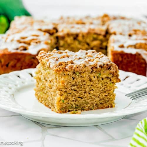 A square slice of Eggless Zucchini Coffee Cake sits on a white plate, with the remaining cake and a green striped napkin visible in the background.