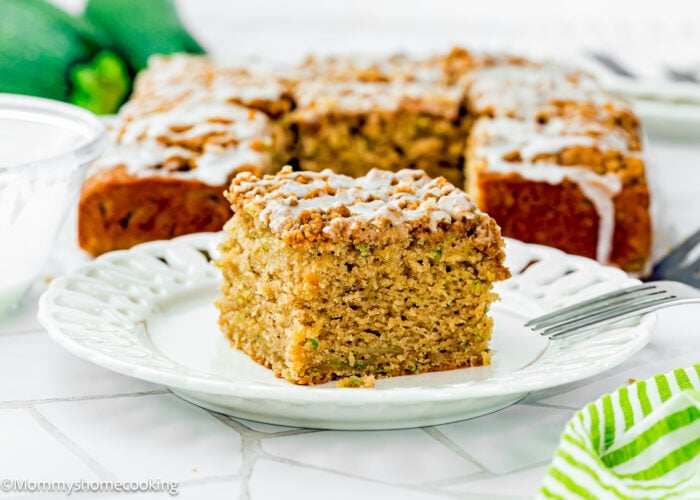 A square slice of Eggless Zucchini Coffee Cake sits on a white plate, with the remaining cake and a green striped napkin visible in the background.