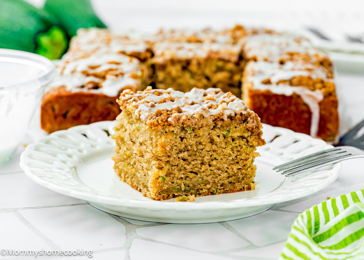 A square slice of Eggless Zucchini Coffee Cake sits on a white plate, with the remaining cake and a green striped napkin visible in the background.