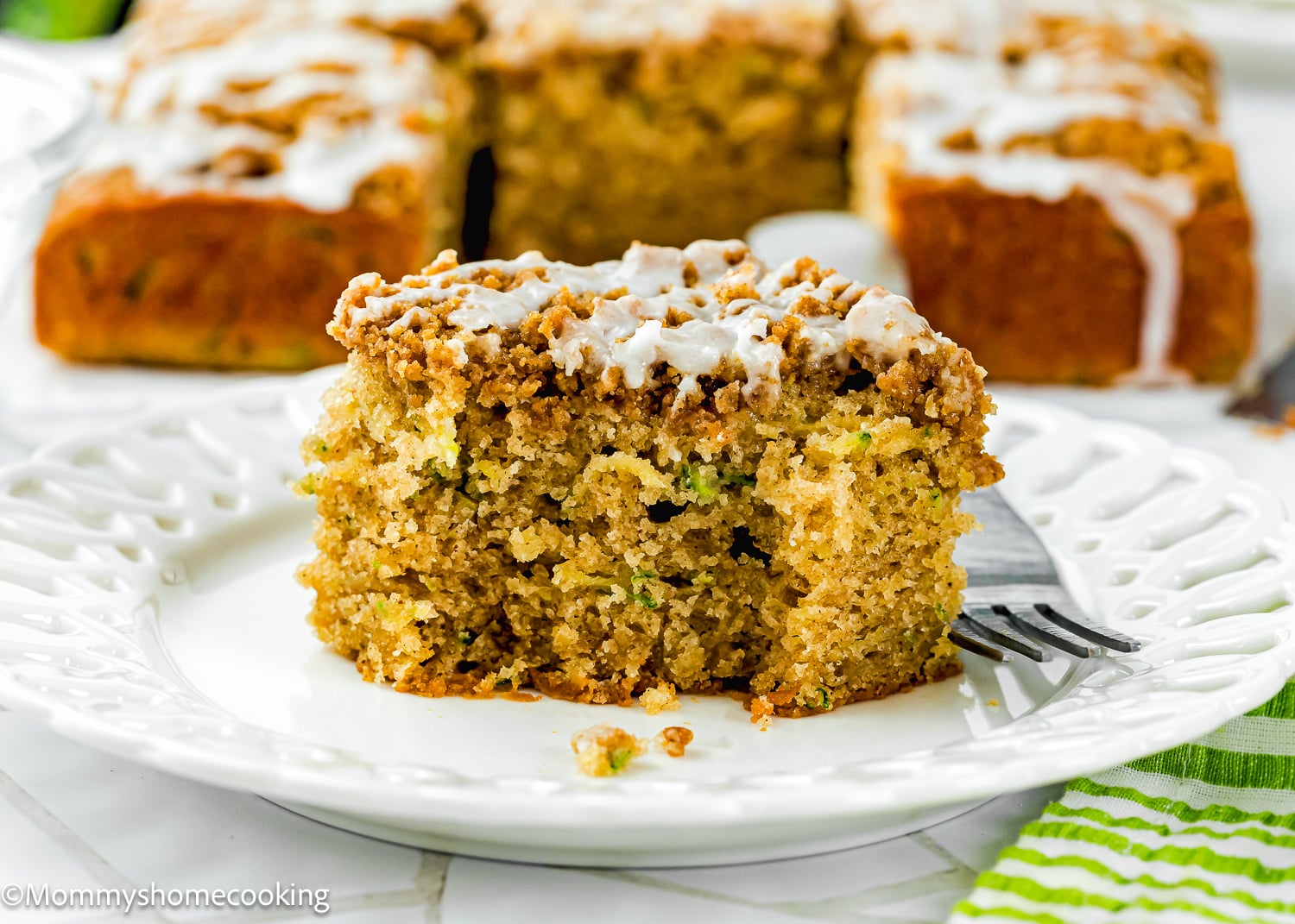 A slice of crumb-topped Eggless Zucchini Coffee Cake with icing sits on a white plate, accompanied by a fork and more cake pieces in the background.