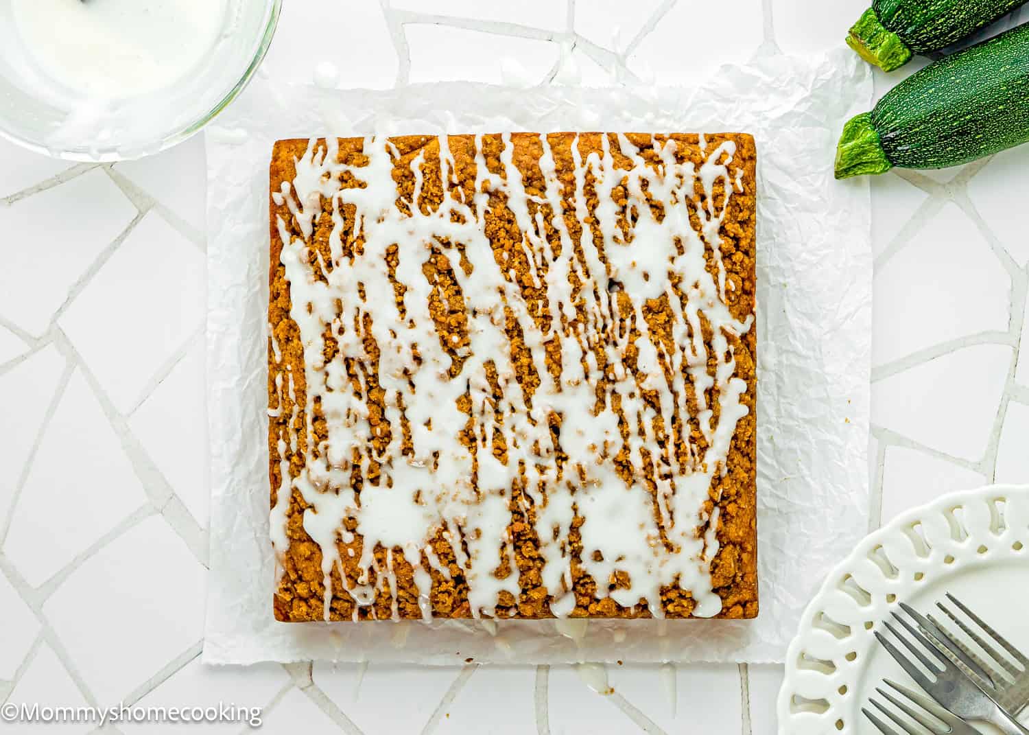 A square Eggless Zucchini Coffee Cake with white icing drizzle sits on parchment paper next to two fresh zucchinis and a white plate on a tiled surface.