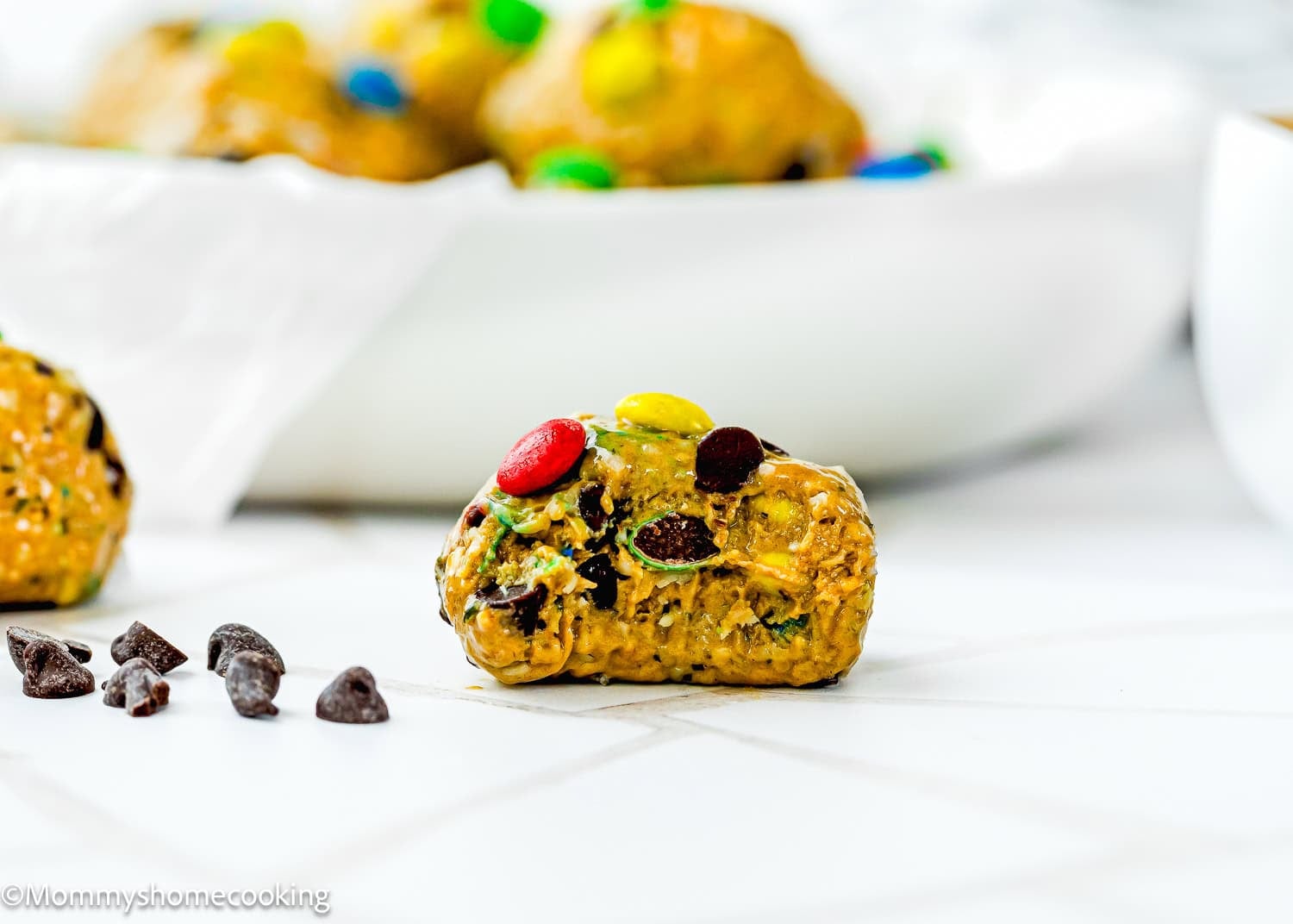 A close-up of a Peanut-Free Monster Cookie Protein Energy Bite with visible chocolate chips and colorful candy-coated chocolates on a white surface.