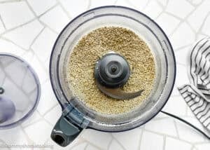 Top view of a food processor containing ground oats on a white tiled surface, with the lid off and a striped cloth to the side—perfect for making Peanut-Free Monster Cookie Protein Energy Bites.