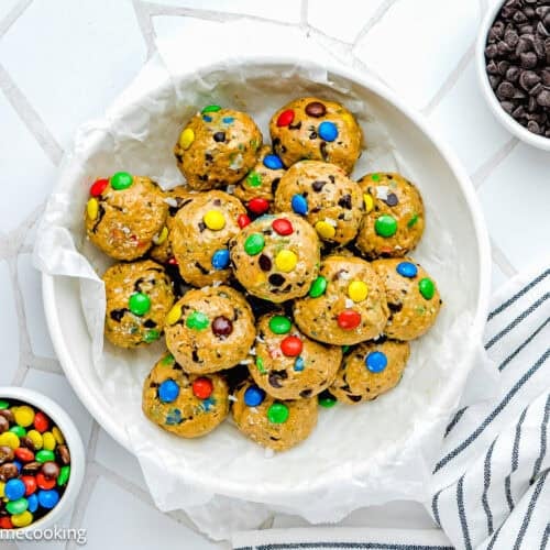 A white bowl filled with Peanut-Free Monster Cookie Protein Energy Bites with colorful candies, next to bowls of mini chocolate chips and candies, on a white surface with a striped towel.