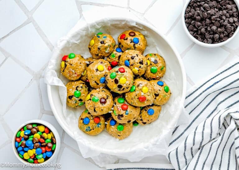 A white bowl filled with Peanut-Free Monster Cookie Protein Energy Bites with colorful candies, next to bowls of mini chocolate chips and candies, on a white surface with a striped towel.