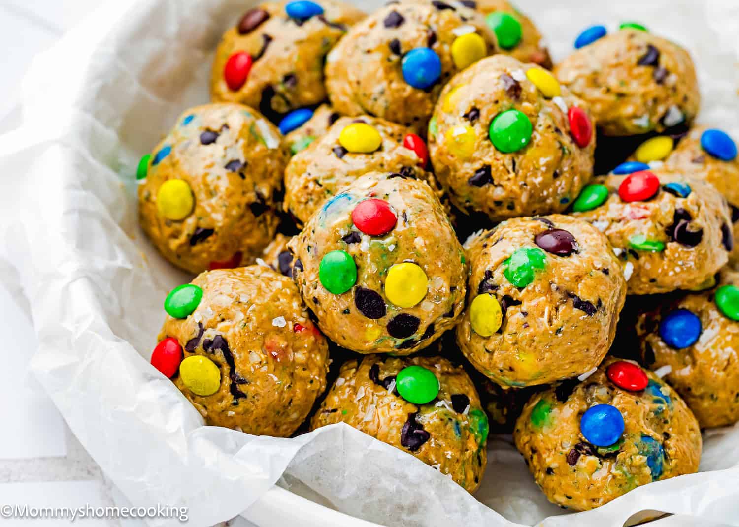 A bowl of Peanut-Free Monster Cookie Protein Energy Bites with chocolate chips and colorful candy-coated chocolates on parchment paper.