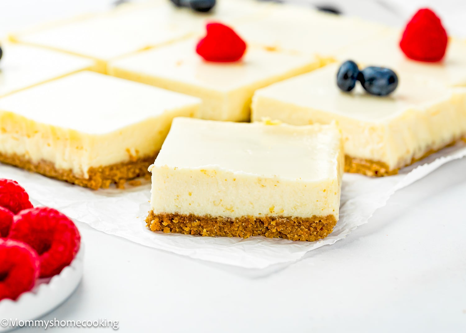 Eggless Cheesecake Bars with a graham cracker crust are arranged on parchment paper, some topped with fresh raspberries and blueberries.