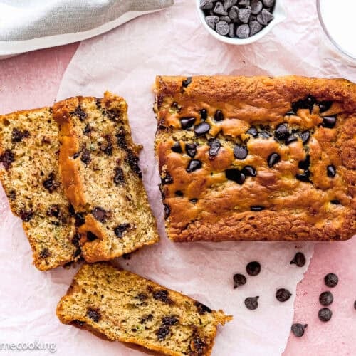 A loaf of chocolate chip banana bread with two slices cut, chocolate chips scattered on parchment, and a glass of milk on a pink surface—perfectly capturing the cozy vibe you’d expect from homemade treats like Eggless Chocolate Chip Zucchini Bread.