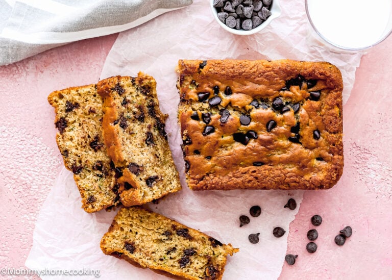 A loaf of chocolate chip banana bread with two slices cut, chocolate chips scattered on parchment, and a glass of milk on a pink surface—perfectly capturing the cozy vibe you’d expect from homemade treats like Eggless Chocolate Chip Zucchini Bread.