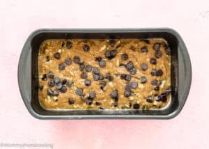A loaf pan filled with unbaked Eggless Chocolate Chip Zucchini Bread batter topped with chocolate chips, set on a light pink surface.