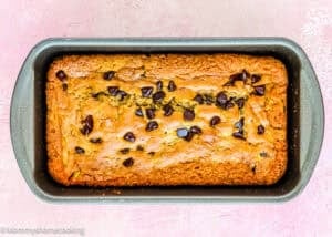 Eggless Chocolate Chip Zucchini Bread baked in a rectangular metal pan, topped with chocolate chips, and placed on a light pink textured surface.