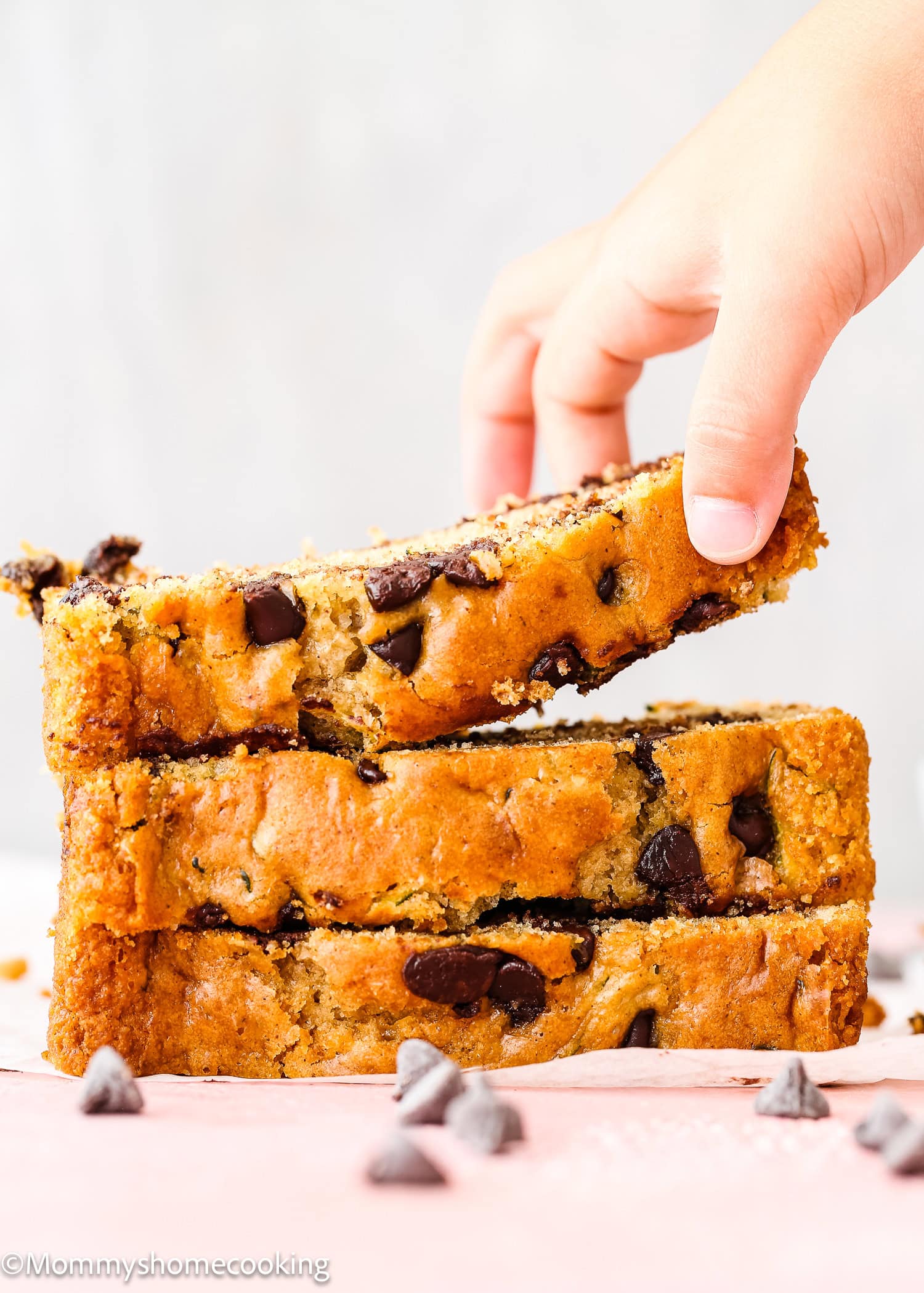 A hand lifting a slice of Eggless Chocolate Chip Zucchini Bread from a stack of three slices on a light surface, with chocolate chips scattered nearby.