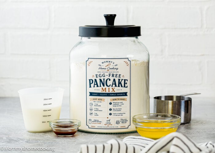 A large jar labeled “Homemade Egg-Free Pancake Mix” sits on a counter with a measuring cup, a bowl of melted butter, a measuring cup of milk, and a small dish of vanilla extract.